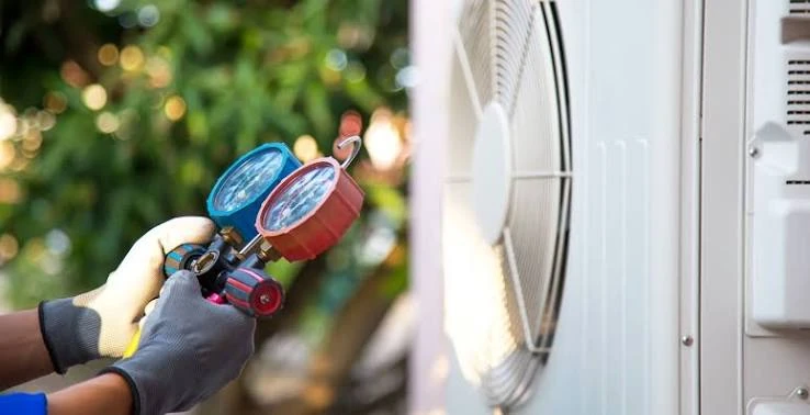 Gloved hands holding refrigerant pressure gauges beside an outdoor cooling unit